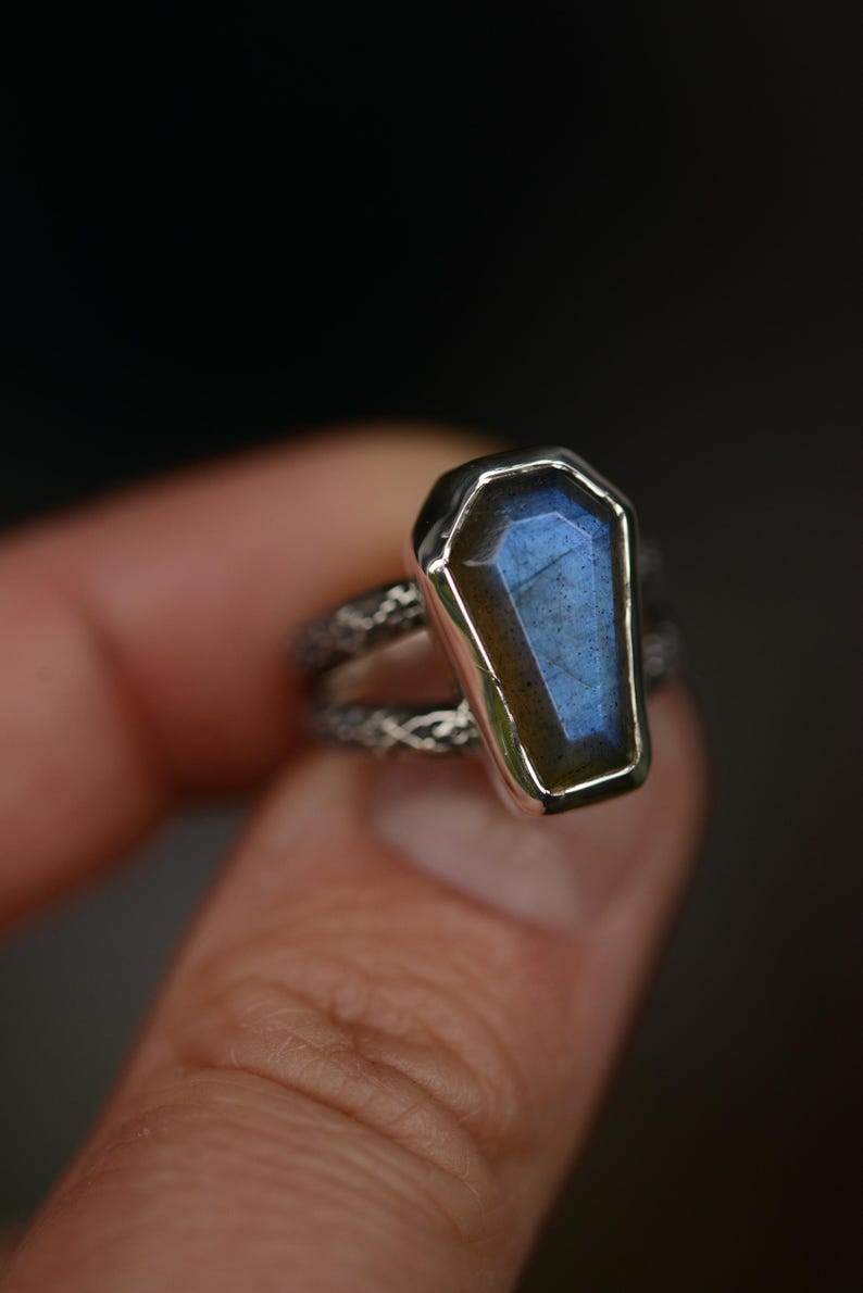 May include: A silver ring featuring a coffin-shaped, blue-hued gemstone. The gemstone is set in a silver bezel, and the band has a textured, patterned design. The ring is held up against a dark background.