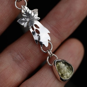 May include: A silver necklace pendant featuring a polished metal ghost shape with a leaf-like top and cut-out facial features, connected by a silver jump ring to a faceted, translucent olive-green gemstone set in a smooth silver bezel frame.