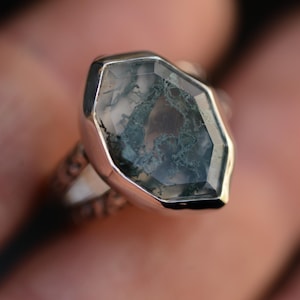 Könnte beinhalten: Ein Silberring mit einem großen, sechseckig geschliffenen Moosachat-Edelstein. Der Stein ist durchscheinend und weist in der Mitte dunkelgrüne, astartige Einschlüsse auf. Der Metallring hat eine texturierte, gemusterte Oberfläche an den Seiten.