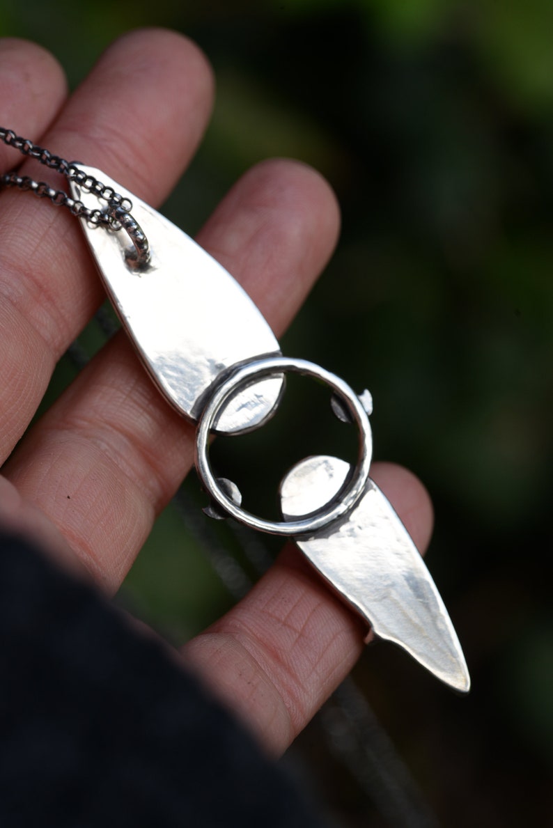 May include: A silver pendant necklace with a unique design featuring a circle with a smaller circle inside. The pendant is attached to a silver chain.