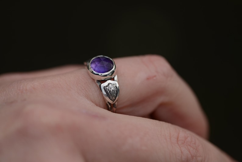 May include: A silver ring with a purple amethyst gemstone set in a textured band. The band features a decorative design with a shield-like shape.