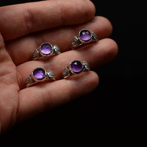 May include: Four silver rings featuring purple gemstones. The rings are set with round cabochon stones and have intricate details on the bands.