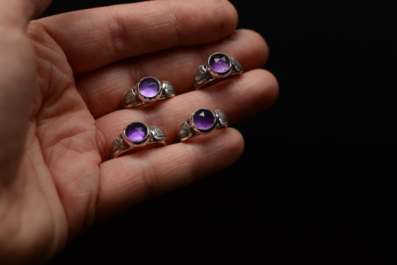 May include: Four silver rings featuring purple gemstones. The rings are set with round cabochon stones and have intricate details on the bands.