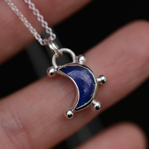 May include: A silver necklace featuring a crescent moon pendant. The pendant is inlaid with a deep blue stone and accented with four small silver spheres along the outer edge. The piece hangs from a delicate silver chain with small circular links.