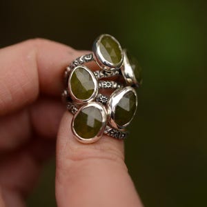 Op de afbeelding: Een verzameling zilveren ringen, elk met een grote, ovaal geslepen, olijfgroene edelsteen. De ringen zijn op een vinger gestapeld en tonen hun ingewikkelde, bloemmotiefbanden. De edelstenen hebben een facetgeslepen oppervlak.