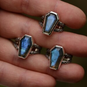 May include: Silver rings with coffin-shaped, iridescent blue gemstones. The rings have a textured band. The gemstones have a silver border. The rings are displayed against a blurred green background.