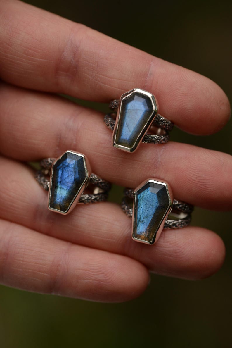 May include: Silver rings with coffin-shaped, iridescent blue gemstones. The rings have a textured band. The gemstones have a silver border. The rings are displayed against a blurred green background.