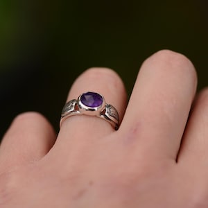 May include: A silver ring featuring a purple amethyst gemstone. The ring is worn on a finger.