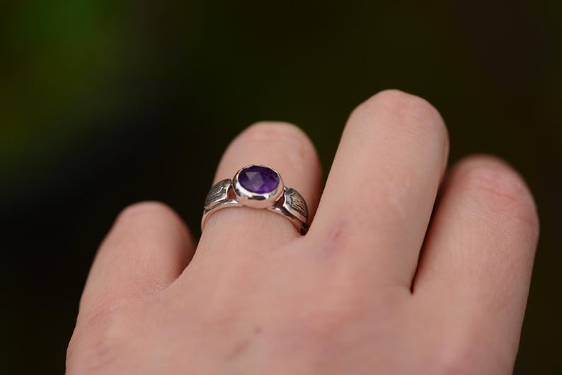 May include: A silver ring featuring a purple amethyst gemstone. The ring is worn on a finger.