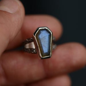 May include: A silver ring featuring a coffin-shaped labradorite stone. The stone has a blue and green iridescent sheen. The ring's band has a textured, ornate design. The ring is held between fingers against a dark background.