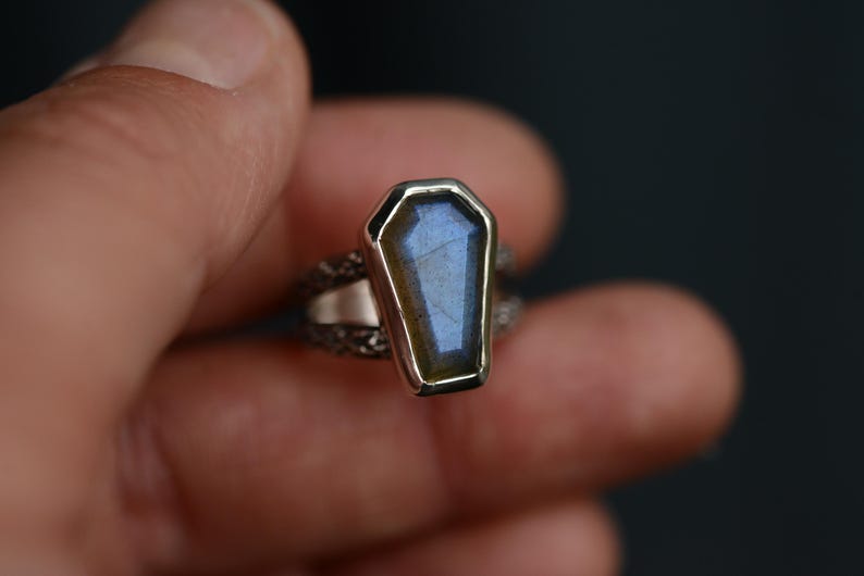 May include: A silver ring featuring a coffin-shaped labradorite stone. The stone has a blue and green iridescent sheen. The ring's band has a textured, ornate design. The ring is held between fingers against a dark background.