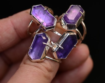 May include: A collection of four silver rings, each featuring a faceted, elongated hexagonal purple gemstone set in a prong setting. The stones exhibit varying shades of translucent violet and deep purple, secured by metallic prongs on thin bands.