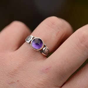 May include: A silver ring with a purple amethyst gemstone set in a bezel setting. The ring has a textured band.