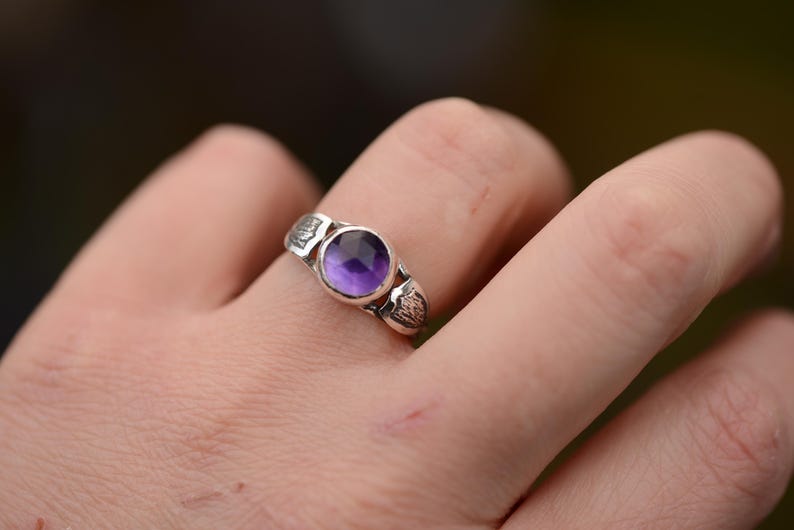 May include: A silver ring with a purple amethyst gemstone set in a bezel setting. The ring has a textured band.