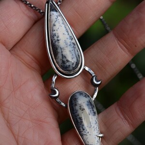 May include: A silver necklace with a teardrop-shaped pendant featuring a white and black dendritic agate stone. The pendant is attached to a silver chain with two crescent moon charms.