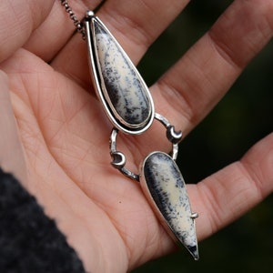 May include: A silver necklace with two teardrop-shaped pendants featuring a white and black dendritic agate stone.