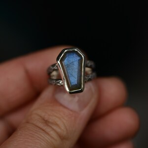 May include: A silver ring featuring a coffin-shaped, blue-hued gemstone. The gemstone is set in a silver bezel, and the band has a textured, twisted design. The ring is held in a person's hand against a dark background.