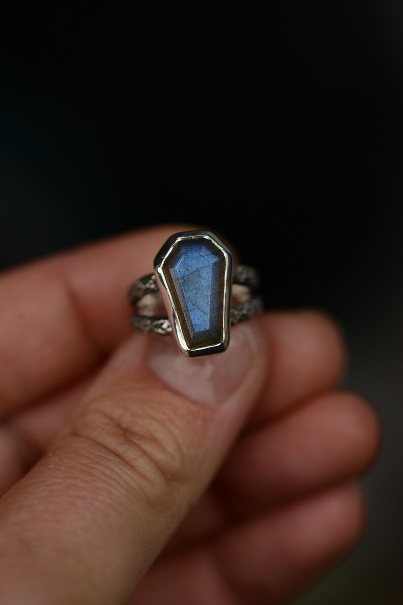 May include: A silver ring featuring a coffin-shaped, blue-hued gemstone. The gemstone is set in a silver bezel, and the band has a textured, twisted design. The ring is held in a person's hand against a dark background.
