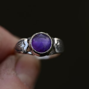 May include: A silver ring featuring an intricate design and a round, purple amethyst gemstone.