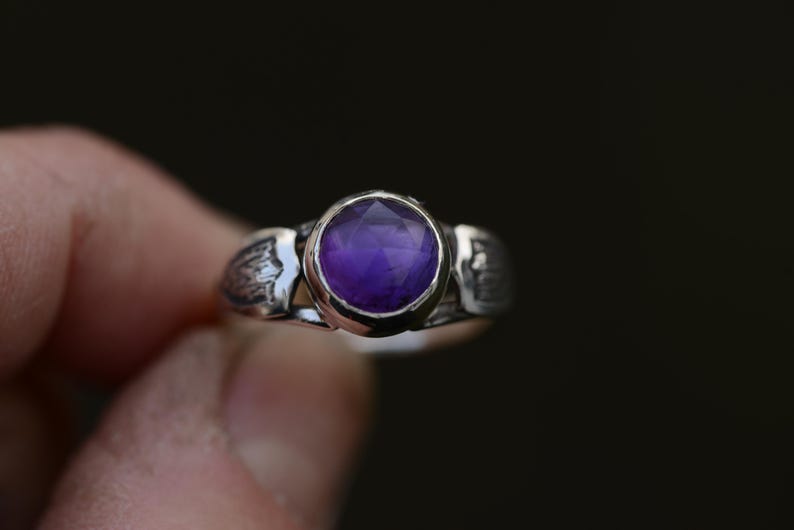 May include: A silver ring featuring an intricate design and a round, purple amethyst gemstone.