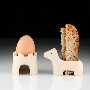 May include: Two ceramic egg cups, one shaped like a castle and the other like a horse, with a boiled egg in the castle cup and two slices of bread in the horse cup.