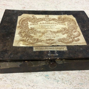 May include: A vintage, rectangular box with a dark, textured surface. A faded label with ornate script and floral designs is affixed to the top. The label reads "BELS AROMATIQUES COMPRIMÉS pour le Bain" and "MORNY 201, Regent Street LONDON". A small metal plate reads "SPECIALLY PREPARED FOR EXPORT".