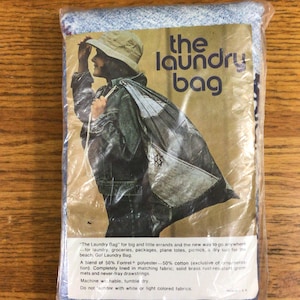 Puede incluir: Una bolsa de lavandería de mezclilla azul con cierre de cordón blanco. La bolsa se muestra en un paquete de plástico con una foto de una persona llevando la bolsa. El texto "the laundry bag" está impreso en el paquete.