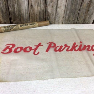 May include: A vintage "Boot Parking" sign in red script lettering on a translucent sheet. A rolled-up brown paper mat labeled "Boot-Mat" rests above the sign against a weathered wood background.