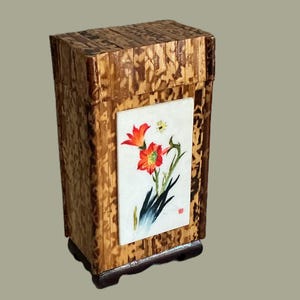 May include: A rectangular wooden box with a lid, featuring a floral painting on the front. The box is constructed from narrow, vertical strips of wood, and the painting depicts orange flowers with green stems and leaves.