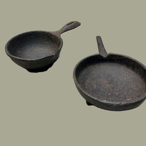May include: Two small, cast iron pans. One is a small skillet with a handle, and the other is a small ladle with a handle. Both are dark gray and appear to be well-used. The pans are on a light green background.