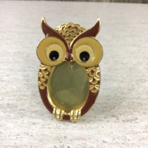 May include: An owl-shaped picture frame with a brown, gold, and cream color scheme. The frame has large, round eyes and a detailed feather design. The frame is made of metal and has a small, oval opening for a photo.