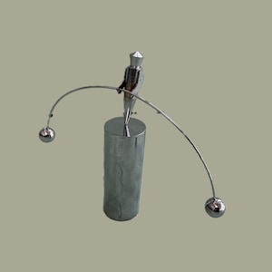 Puede incluir: Una escultura cinética plateada de una figura equilibrándose sobre un cilindro. La figura sostiene una barra curva con una esfera en cada extremo. La escultura está sobre un fondo neutro.