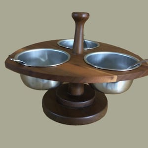 May include: A wooden condiment holder with three stainless steel bowls and a central wooden handle. The holder has a round base and a slightly oval top.