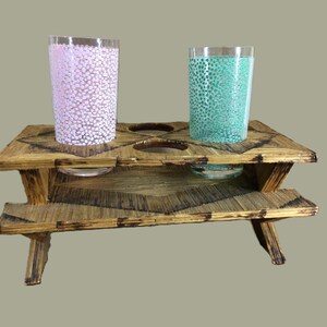 May include: A wooden picnic table made of matchsticks with two glass cups with pink and green patterns on top.