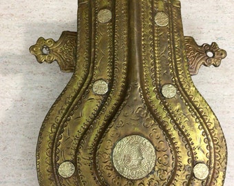 Puede incluir: Una cuerno de pólvora de metal dorado con intrincados diseños grabados y acentos circulares. El cuerno tiene un lazo en la parte superior y dos lazos decorativos en los lados.