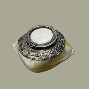 May include: A small square box crafted from iridescent mother-of-pearl panels. The lid features a circular silver-toned metal frame with an intricate embossed scrollwork pattern and a central mother-of-pearl inlay, topped with a small heart-shaped clasp.