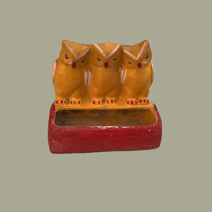 May include: A decorative owl figurine featuring three owls in a row, painted in shades of yellow and white, with red accents. The owls are perched atop a red rectangular base. The figurine is likely made of ceramic or a similar material.