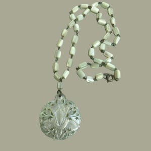 May include: A vintage necklace featuring a carved pendant with a floral design, suspended from a strand of off-white, rectangular beads. The necklace has a silver-tone clasp and is displayed against a neutral background.