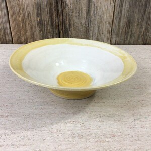 Peut inclure: Un bol en céramique blanche avec un bord jaune et un motif en spirale jaune au centre. Le bol est posé sur une surface brun clair.