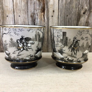 Puede incluir: Dos macetas de cerámica en blanco y negro con un borde dorado. Cada maceta presenta una ilustración en blanco y negro de un caballero a caballo atravesando un bosque.