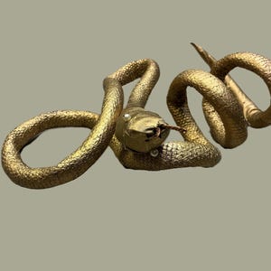 May include: A golden-colored snake sculpture with a textured surface. The snake is coiled and has a detailed head with small pearl accents. The sculpture is set against a neutral background.