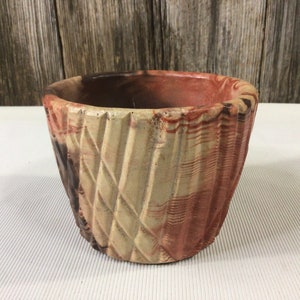 May include: A small, hand-crafted ceramic planter with a textured, geometric design. The planter is glazed in shades of brown, red, and cream.