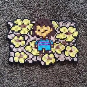 May include: A pixel art bead sprite of a character from the video game Undertale, surrounded by yellow flowers. The character is wearing a blue and purple striped shirt and brown hair.
