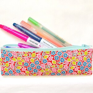 May include: A red and white floral pencil case with a light blue zipper. The case is filled with various pens and pencils, including a pink highlighter, a blue pen, and a green pen.