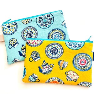 May include: Two zippered pouches with a blue and yellow floral pattern. The pouches feature a repeating design of teapots and plates with floral patterns.