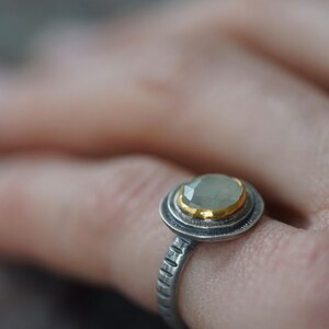 May include: A close-up of a silver ring with a light green gemstone and gold accents. The ring has a textured band and a halo design around the gemstone. The ring is on a finger.