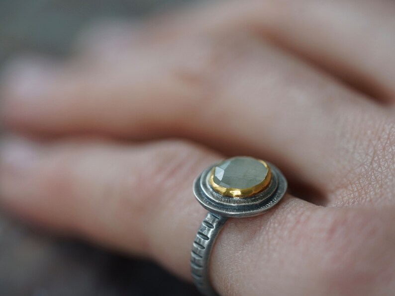 May include: A close-up of a silver ring with a light green gemstone and gold accents. The ring has a textured band and a halo design around the gemstone. The ring is on a finger.