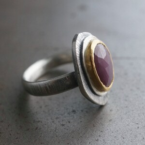 May include: A silver ring with a textured band and a gold-plated setting. The ring features a large, oval-shaped, faceted, deep red gemstone. The setting is oval and layered, adding dimension to the design.