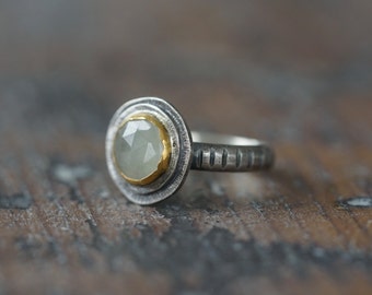 May include: A silver ring with a pale green, faceted gemstone set in a gold bezel. The ring has a textured band with vertical lines. The gemstone is round and set in a circular setting.