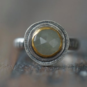 May include: A silver ring with a faceted, pale green gemstone set in a gold bezel. The ring has a textured silver band and a double-layered, textured silver setting around the gemstone.
